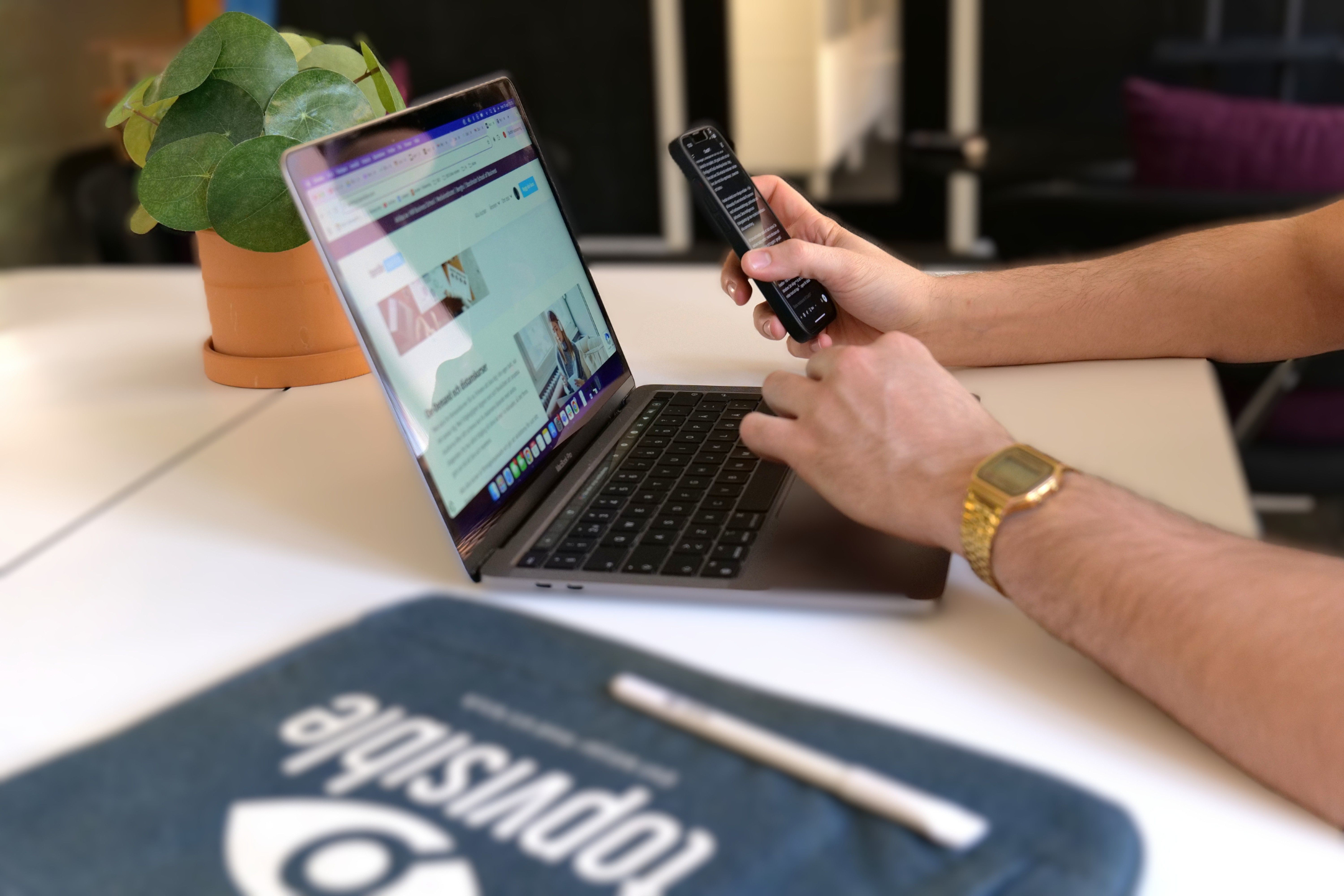 Person som bläddrar i en kurs om digital marknadsföring på en bärbar dator medan hen håller i en smarttelefon, sittande vid en modern arbetsplats med en krukväxt och Topvisible-material på skrivbordet.